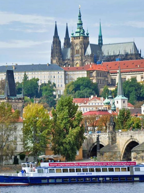 Panoramic view of Prague Castle and Charles Bridge with a cruise boat on the Vltava River.
