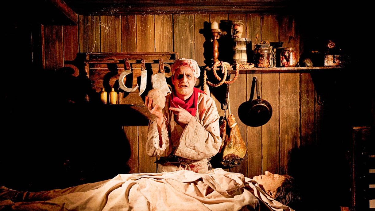 Actor in medieval setting at Amsterdam Dungeon, holding a skull, with tools and props in background.