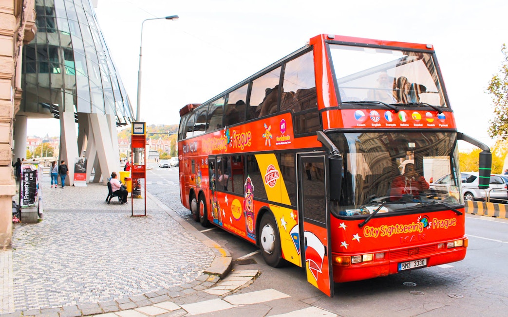 Red City Sightseeing Prague bus near modern architecture on a cobblestone street.
