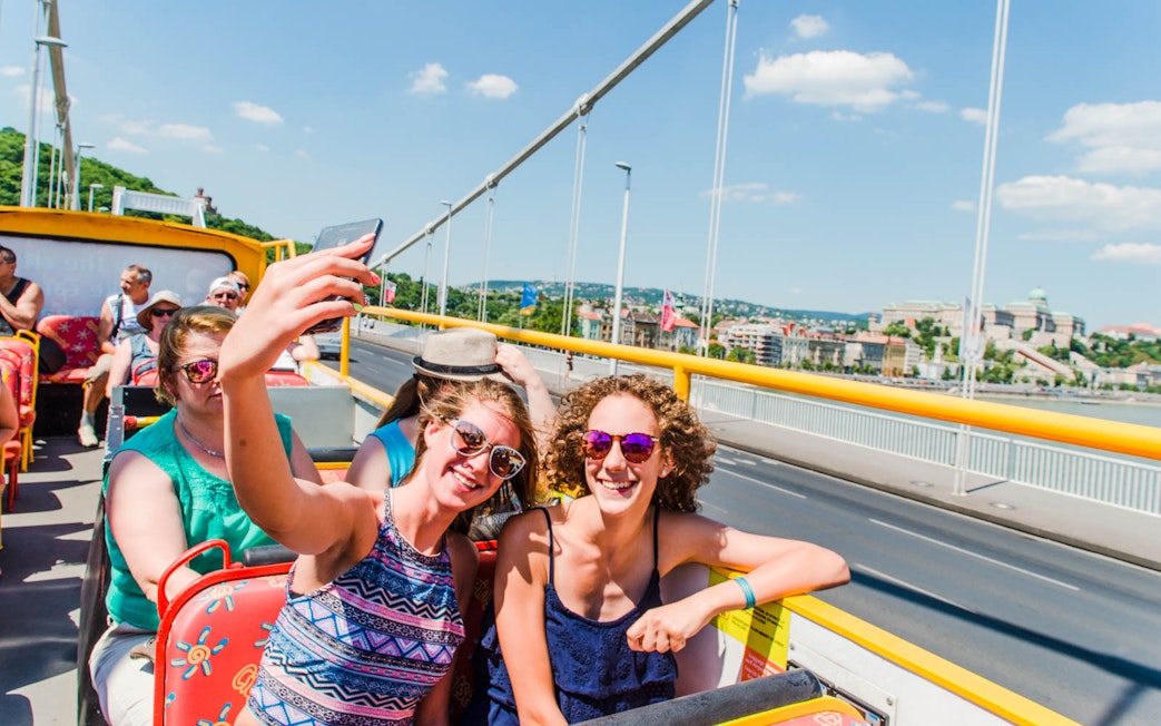 Tourists enjoying a city sightseeing hop-on hop-off bus tour with views of a bridge and cityscape.