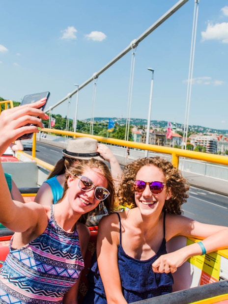 Tourists enjoying a city sightseeing hop-on hop-off bus tour with views of a bridge and cityscape.