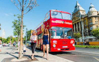 Red City Sightseeing bus in Budapest with tourists walking nearby, Ferris wheel in background.