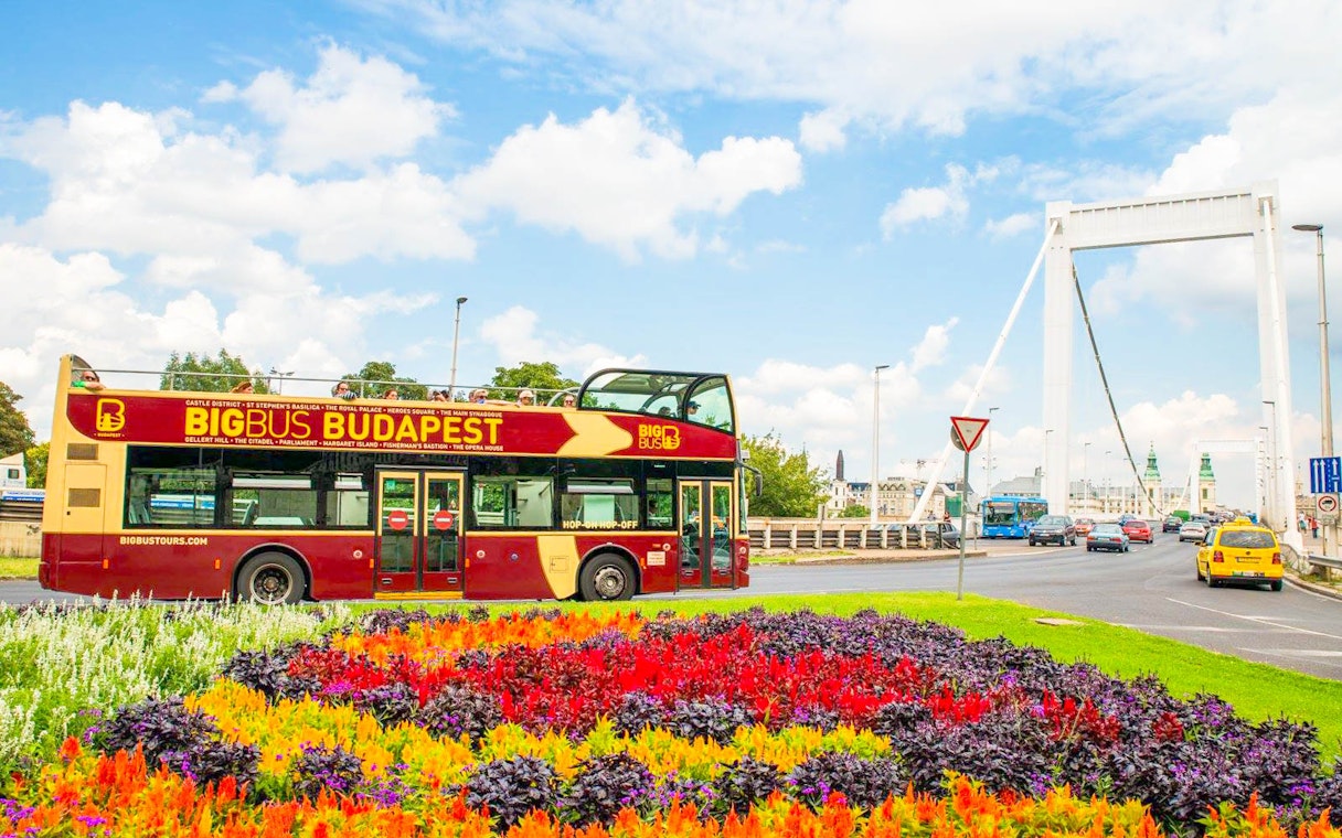 Budapest Big Bus passing by colorful flowers near a bridge.