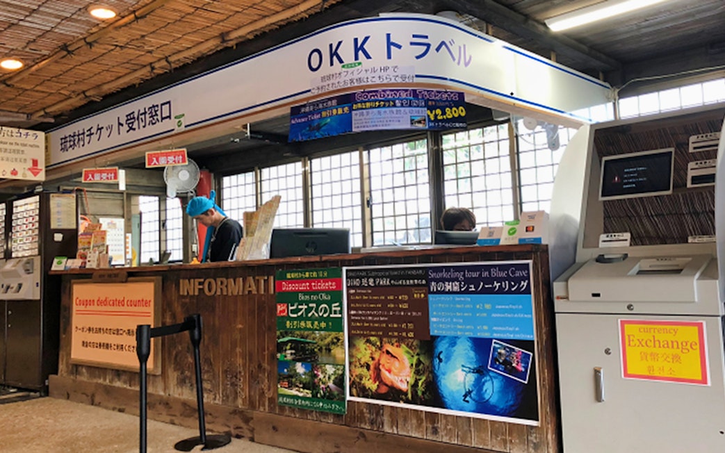 Ticket counter at Okinawa Ryukyu Mura Village with tour and currency exchange information.