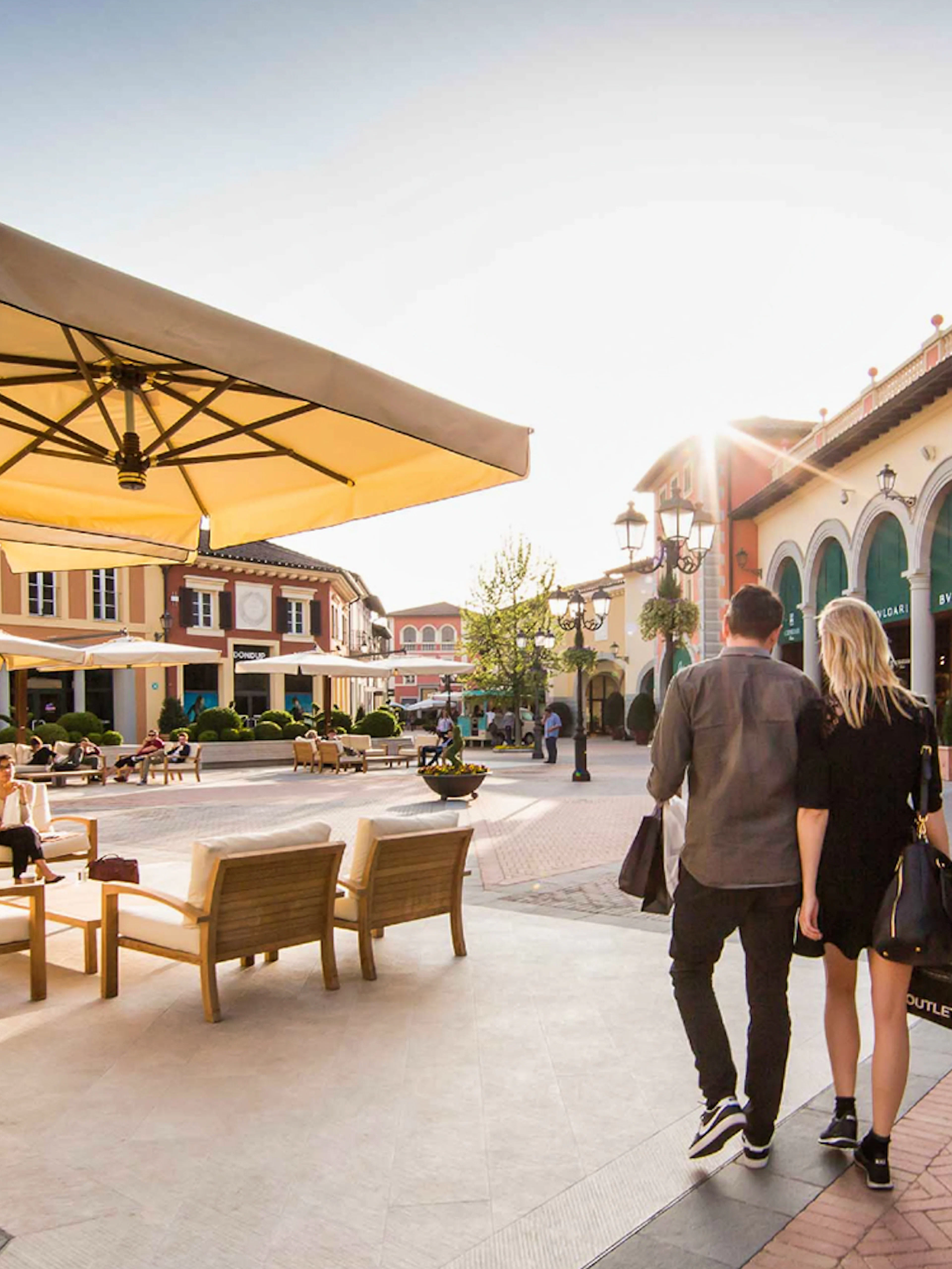 Couple walking at Serravalle Designer Outlet with shops and seating area.