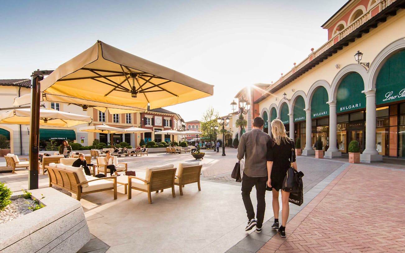 Couple walking at Serravalle Designer Outlet with shops and seating area.