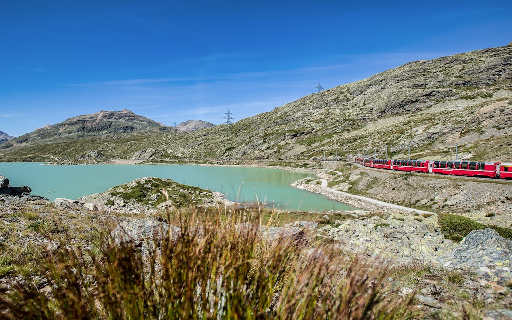 Bernina Express train passing by a turquoise lake in the Swiss Alps on the way to St. Moritz.