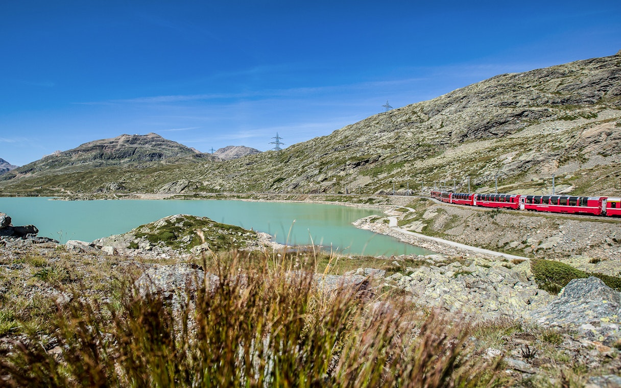 Bernina Express train passing by a turquoise lake in the Swiss Alps on the way to St. Moritz.