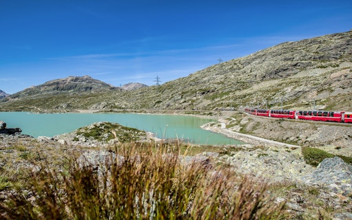 Bernina Express train passing by a turquoise lake in the Swiss Alps on the way to St. Moritz.