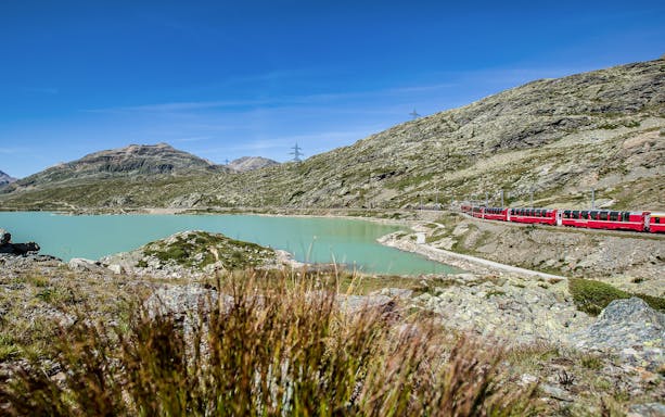 Bernina Express train passing by a turquoise lake in the Swiss Alps on the way to St. Moritz.