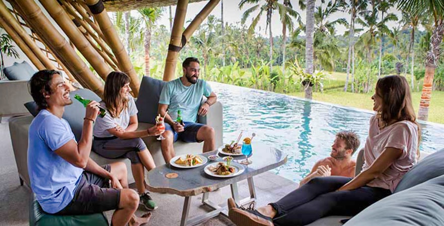 Group relaxing by pool with drinks and food, surrounded by tropical jungle setting.