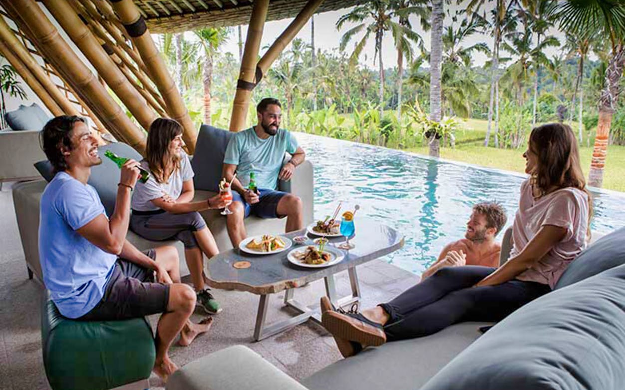 Group relaxing by pool with drinks and food, surrounded by tropical jungle setting.