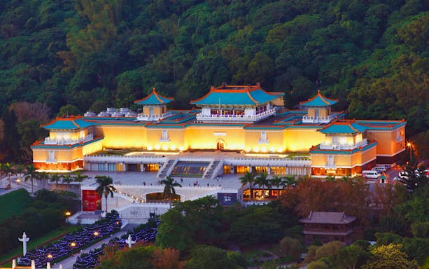 National Palace Museum in Taipei illuminated at dusk, surrounded by lush greenery.