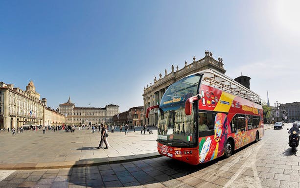 Open-top tour bus in Piazza Castello, Turin, Italy, with historic buildings in the background.
