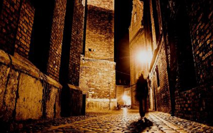 Shadowy figure walking down a dimly lit cobblestone alley in London.