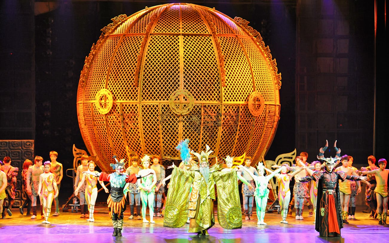 Performers in colorful costumes on stage at Beijing Chaoyang Theatre with a large golden sphere backdrop.