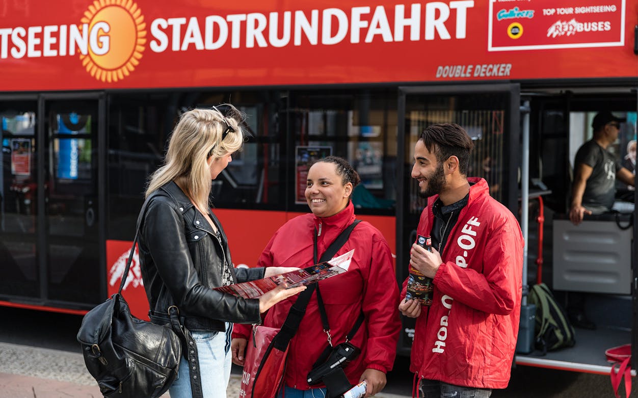 Tourists receiving information from guides in front of a Red Sightseeing Hop On Hop Off bus.