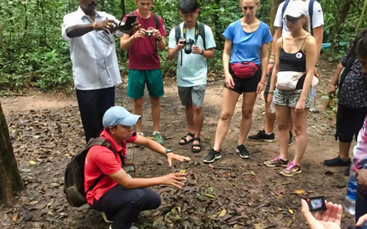 Tour guide explaining Cu Chi Tunnels to visitors in a forest setting.