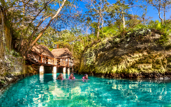 Visitors swimming in a turquoise river surrounded by lush greenery at Xcaret Park, Mexico.