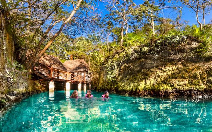 Visitors swimming in a turquoise river surrounded by lush greenery at Xcaret Park, Mexico.