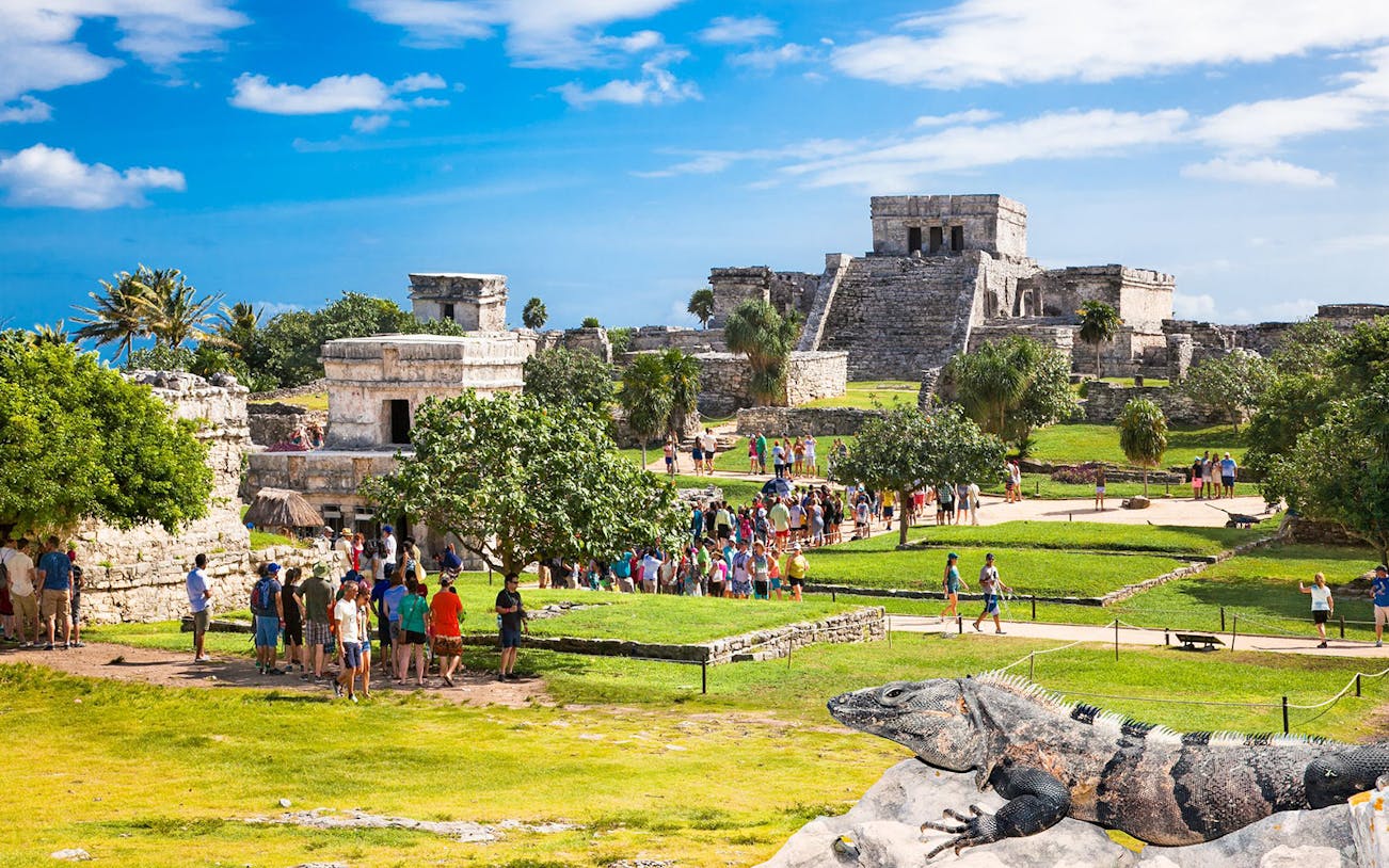 Tulum Ruins with tourists exploring ancient structures and iguana in foreground, Mexico.