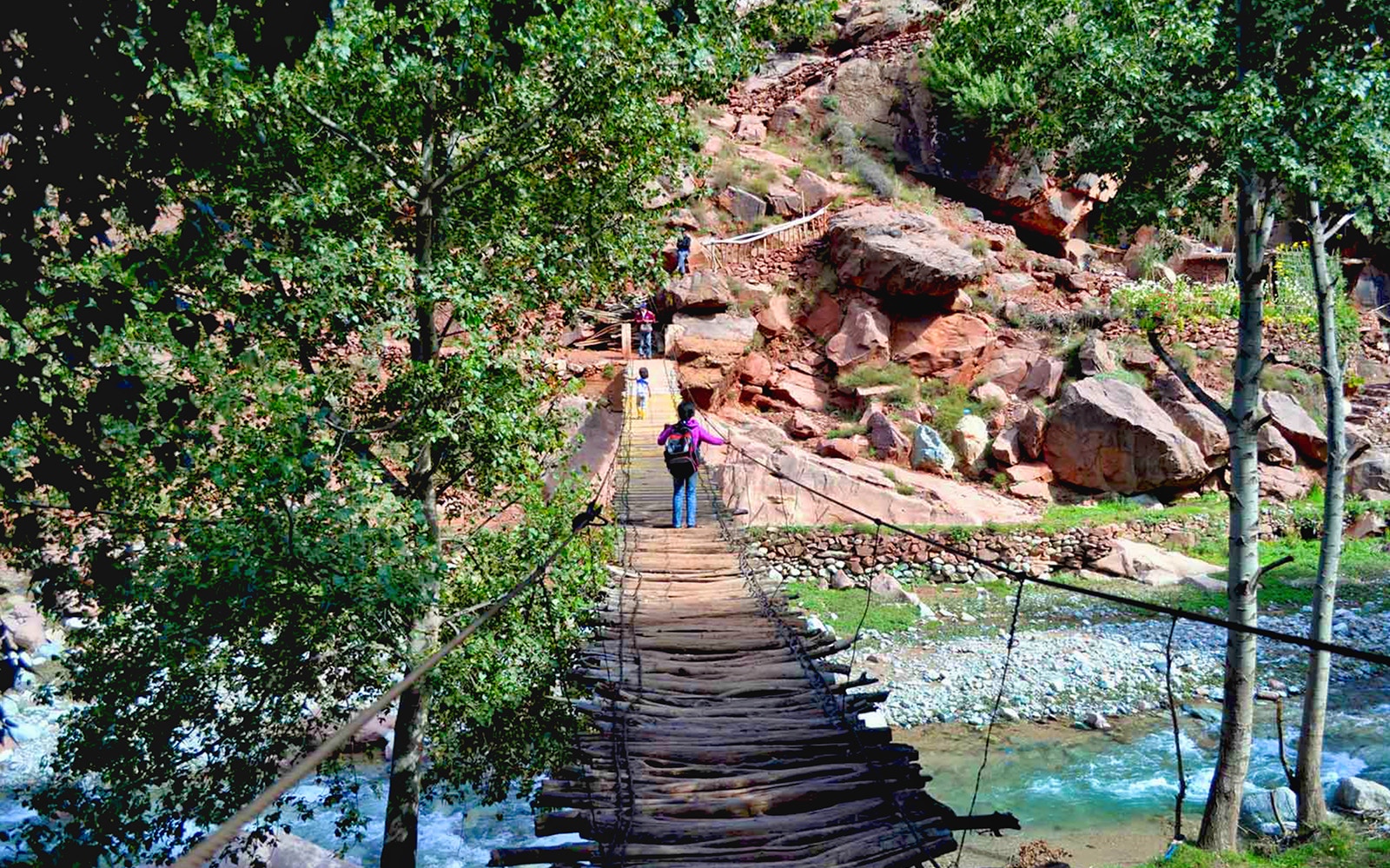 Hikers crossing a wooden bridge in Ourika Valley, Morocco, with rocky landscape.