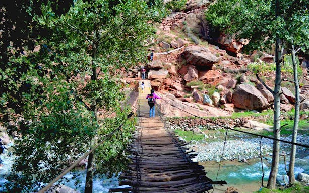 Hikers crossing a wooden bridge in Ourika Valley, Morocco, with rocky terrain and trees.