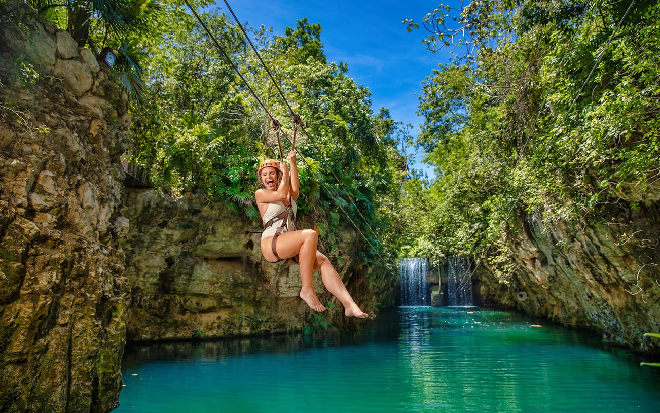 Person ziplining over turquoise water at Xplor Park, surrounded by lush greenery and rocky cliffs.