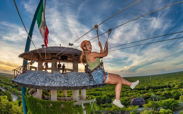 Ziplining at Xplor Park with a view of lush greenery and a thatched-roof platform.