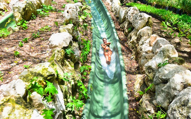 Person sliding down a water slide surrounded by rocks and greenery at Xenses Park.