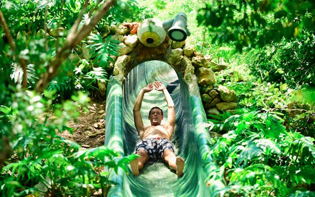 Person sliding down a water slide surrounded by lush greenery at Xenses Park.