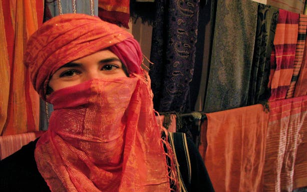 Person in colorful headscarf at Marrakesh souk with hanging textiles in background.