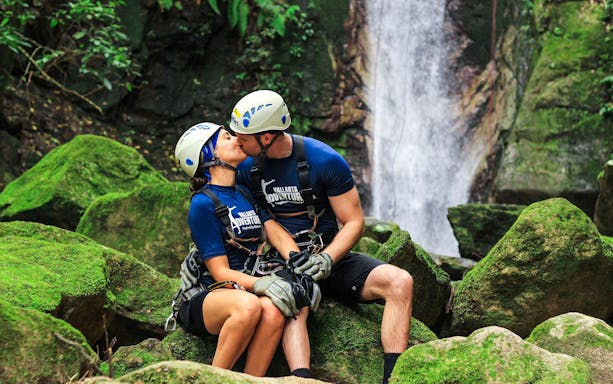 Couple in helmets sitting on mossy rocks near waterfall during Banderas Bay jungle adventure.