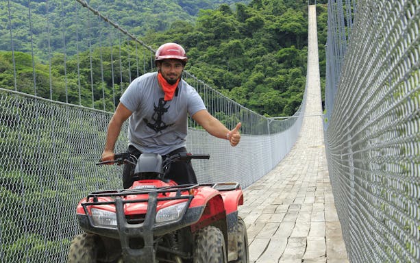 ATV rider crossing Jorullo Suspended Bridge in lush forest setting.