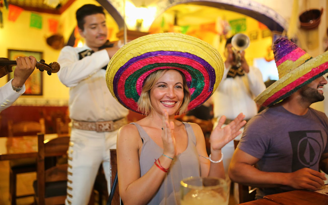 Person enjoying mariachi performance in Cancun restaurant, wearing colorful sombrero.