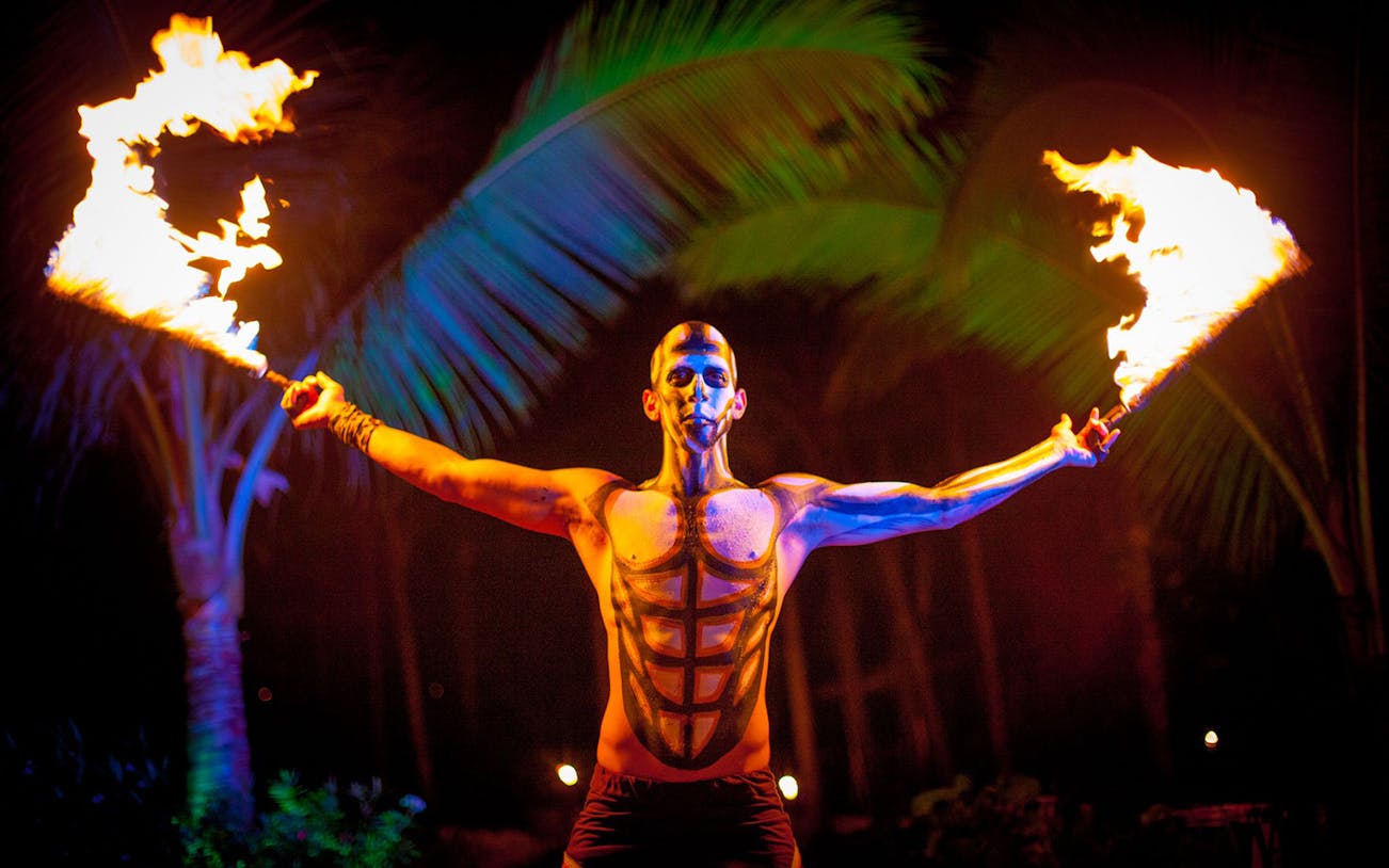 Fire performer at Rhythms of the Night Dinner Show, holding flaming torches with palm trees in the background.