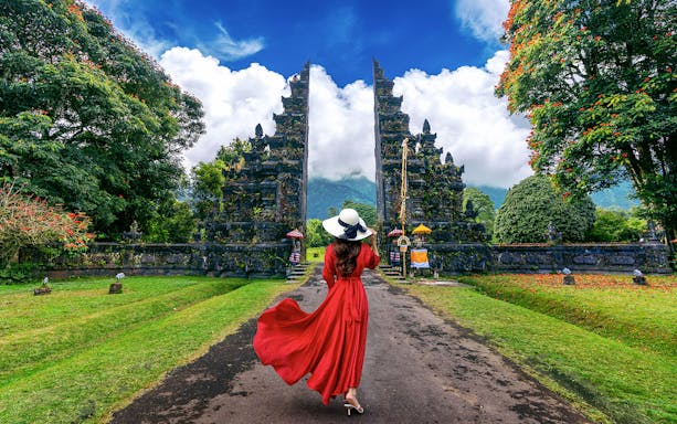 Woman in red dress walking towards Handara Gate in Bali, surrounded by lush greenery.