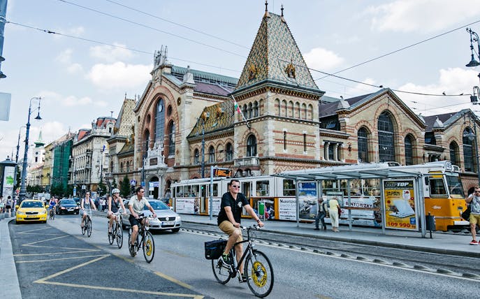 Cyclists riding past the Great Market Hall in Budapest along the Danube River.