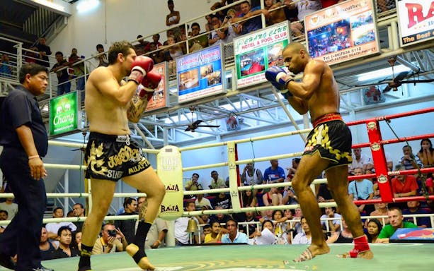 Boxers in a match at Bangla Boxing Stadium, Phuket, with a referee and audience watching.