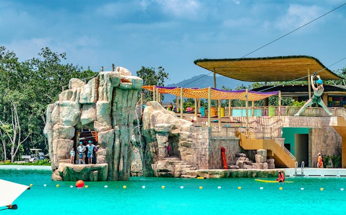 Zipline over turquoise pool at Blue Tree Water Park, Thailand, with rock formations and lounging area.