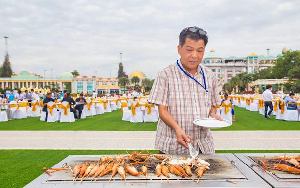 Man grilling seafood at outdoor event, Sukhawadee House, with guests seated in background.