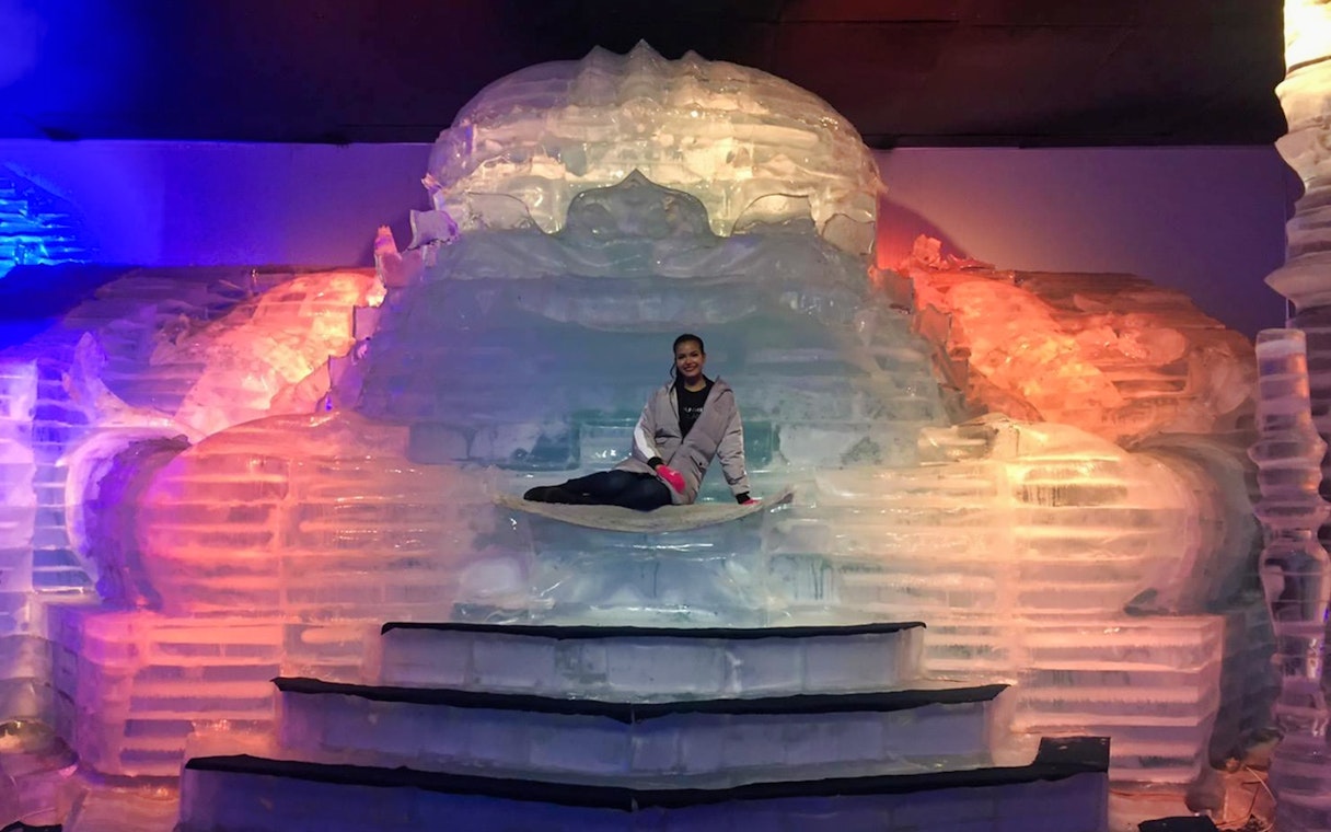 Person sitting on an ice sculpture at Frost Magical Ice of Siam, illuminated by colorful lights.
