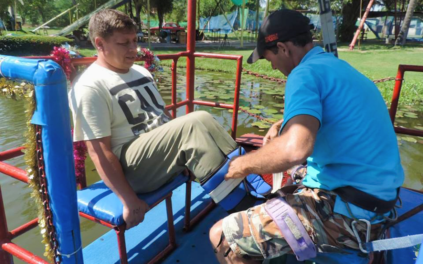 Man preparing for bungy jump with guide securing harness near water.