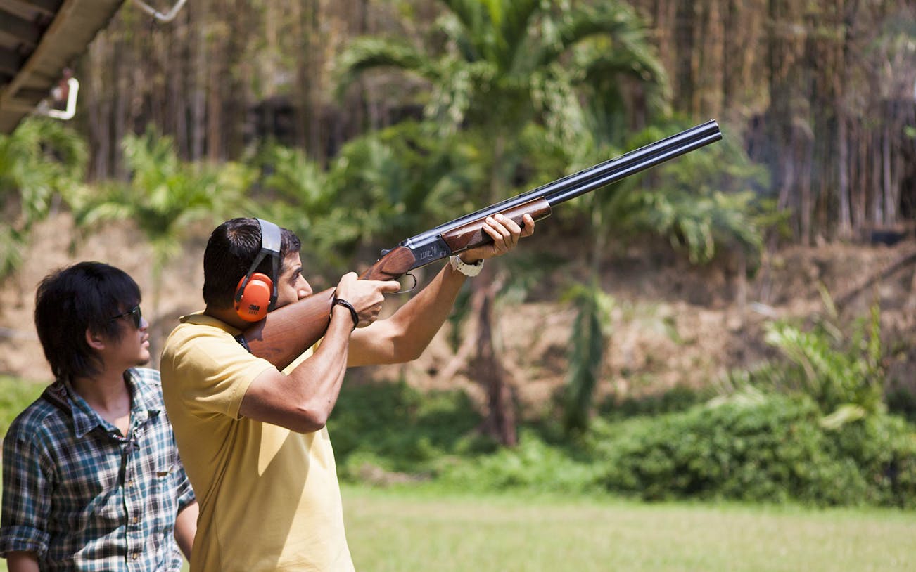 Man aiming a shotgun at Pattaya Shooting Park, Thailand, with an instructor nearby.