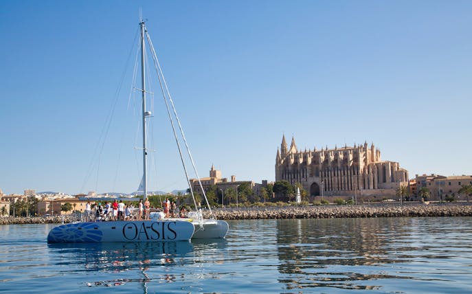 Catamaran sailing near Palma Cathedral, Mallorca, with tourists on deck.