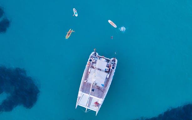 Aerial view of a catamaran with people kayaking and paddleboarding nearby.
