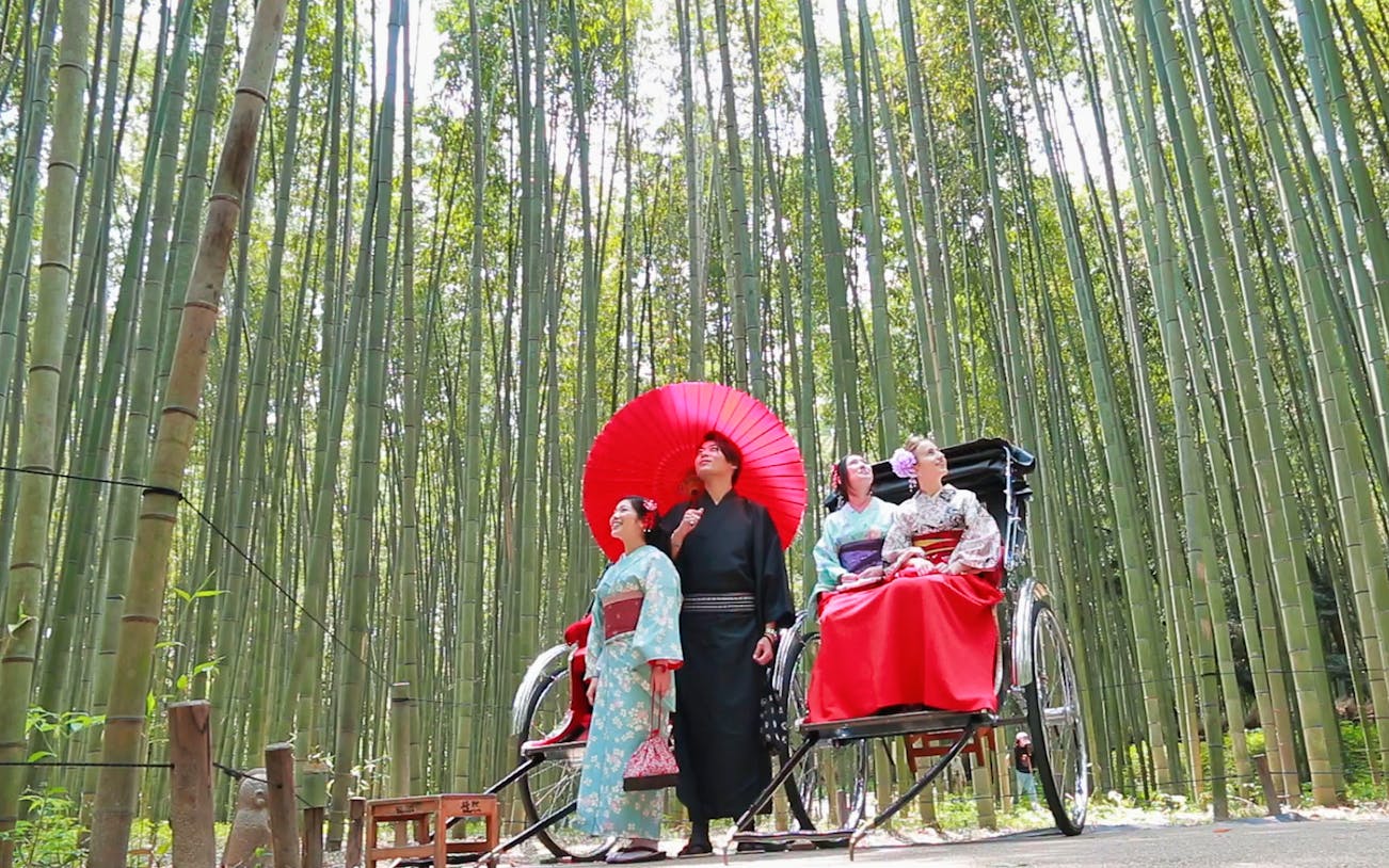People in traditional attire with a red umbrella and rickshaw in Kyoto's bamboo grove.