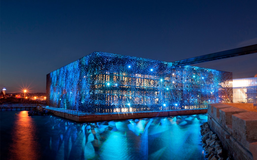 Mucem in Marseille illuminated at night, reflecting on the water.