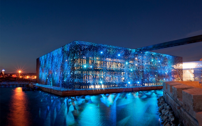 Mucem in Marseille illuminated at night, reflecting on the water.