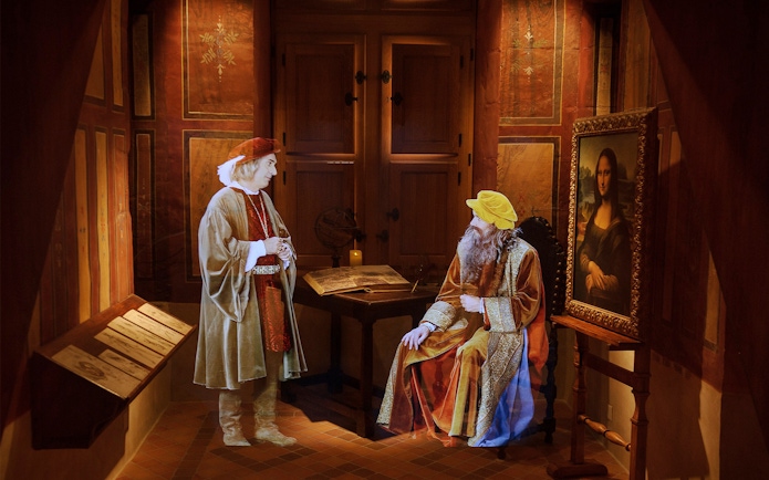 Two historical figures in period clothing at Clos Lucé, with a Mona Lisa replica nearby.